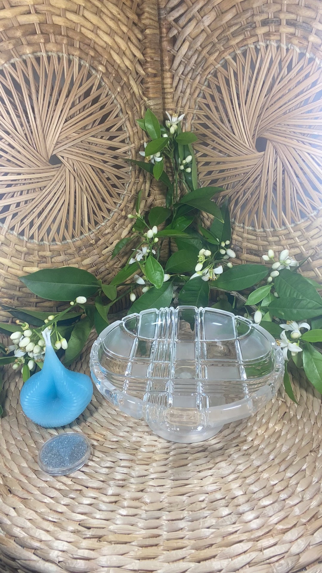 The video shows a blue Destiny Decision Candle with a round coin sized container filled with Destiny Sparkles. There is a fireproof glass plate placed on a natural wicker table and a wicker background. Green leaves and tiny Valencia orange blossoms are in the background.

Demonstrating how to use the Destiny Decision Candle.  Pour the Destiny Sparkles into the glass plate.  Place the candle candle on top of the Destiny Sparkles.  Ask your yes or no question as you light the two wicks.

